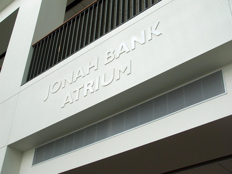 A white concrete wall with the text "JONAH BANK ATRIUM" embossed on it in uppercase letters. The wall has a black horizontal railing near the top.