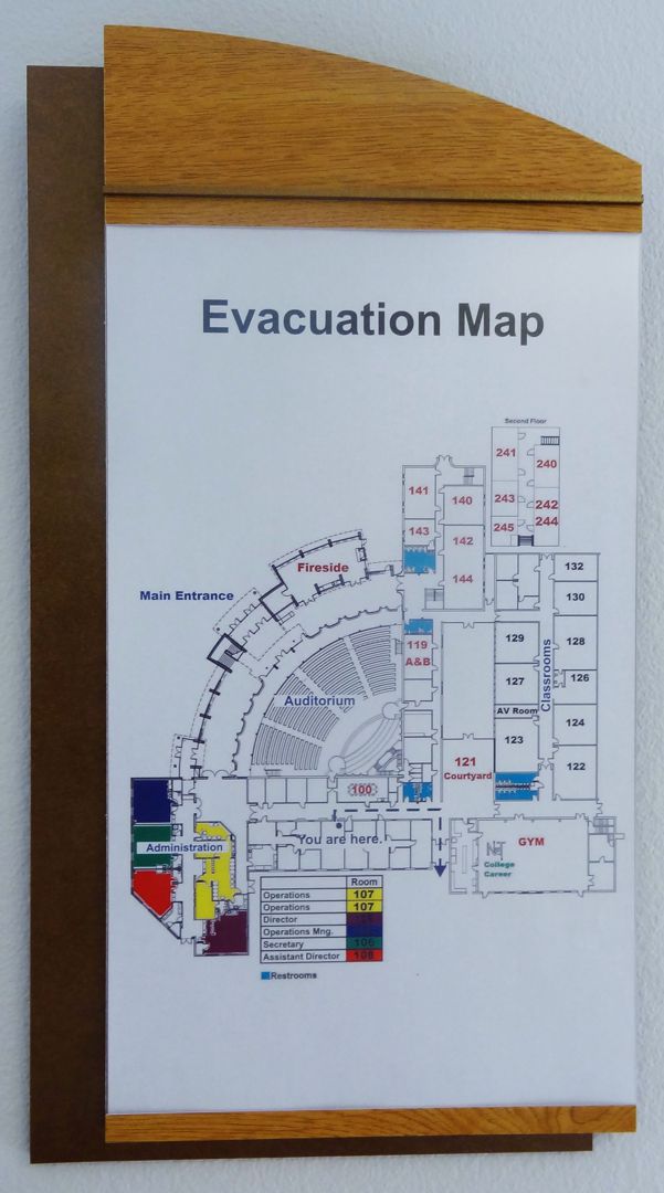A wall-mounted evacuation map framed in wood shows a detailed layout of a building with various rooms numbered. The areas are color-coded, and exits are marked. The map has a title "Evacuation Map" at the top.