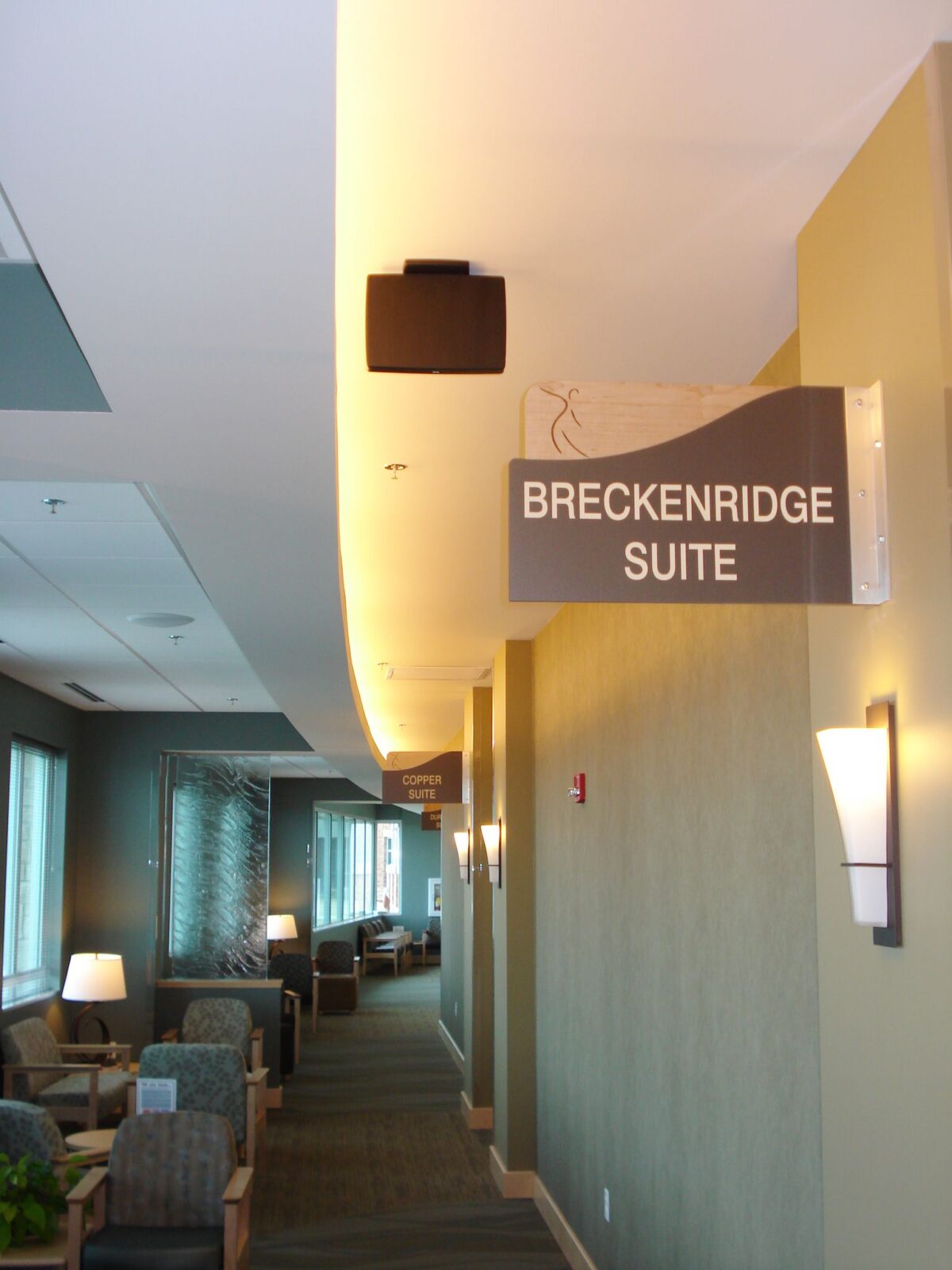A hallway with modern decor and several seating areas lit by floor and wall lamps. A sign hanging from the ceiling reads "BRECKENRIDGE SUITE." Large windows along the left wall let in natural light. The atmosphere appears calm and inviting.