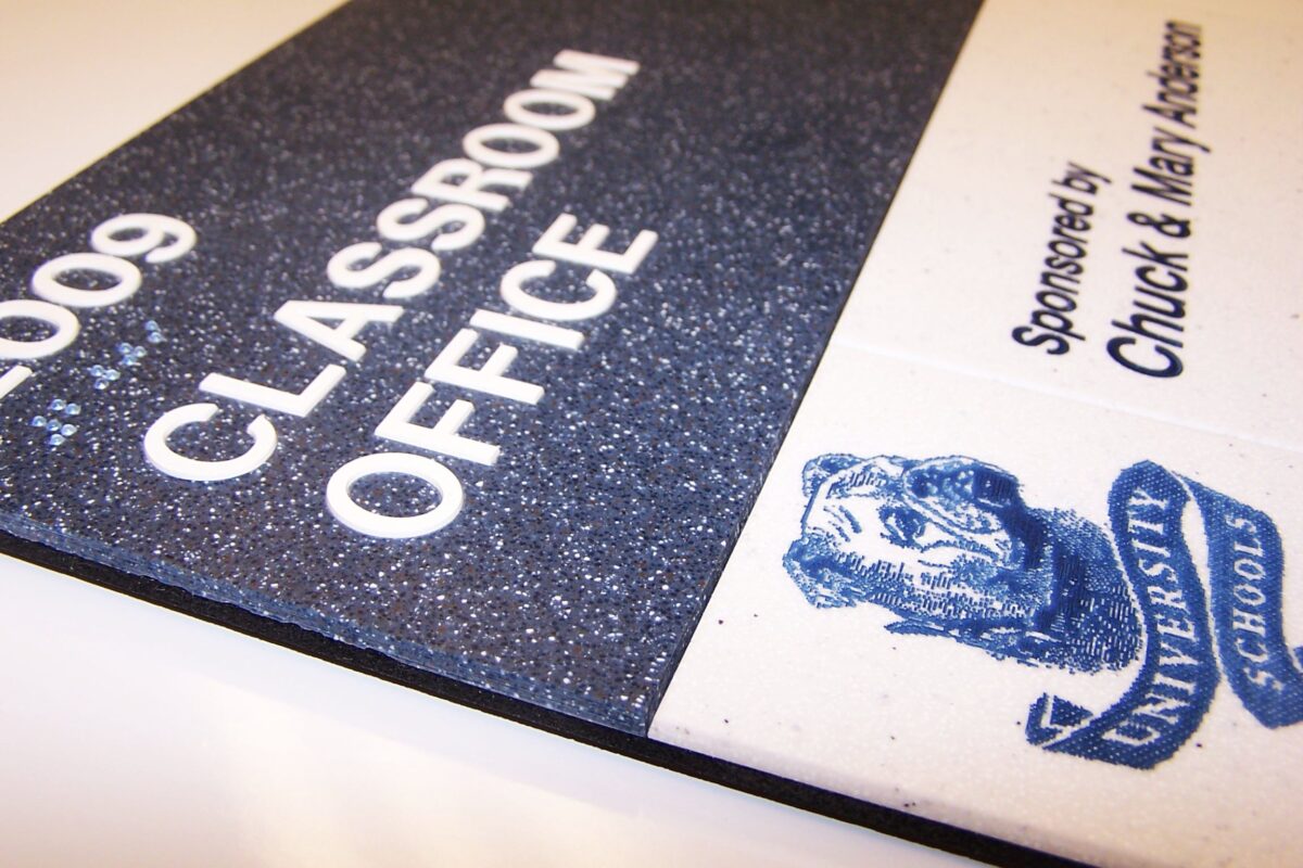 Close-up of a sign indicating "Classroom Office" with the number "2009" on a textured black and white background. The sign is sponsored by "Chuck & Mary Adamson" and features a blue logo with an image of a dog's head and the words "University Schools.
