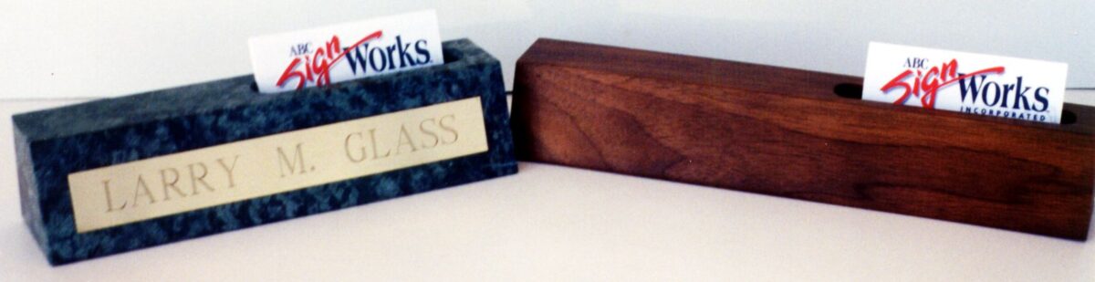 Two nameplate holders are displayed on a surface. The holder on the left is marble with a golden nameplate reading "LARRY M. GLASS." The one on the right is wooden with a slot holding a business card. Both nameplate holders also hold business cards from "The Sign Works.