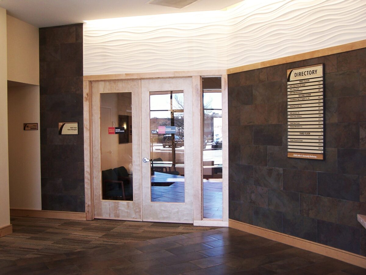A modern lobby with dark tiled walls and floor. Double glass doors in a light wooden frame lead to another room. A directory board is mounted on the right wall, and light fixtures give a soft illumination to the space.