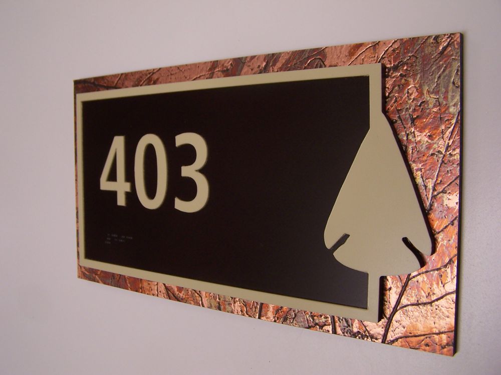 A rectangular sign on a wall displays the number "403" in large beige numerals on a dark brown background. The sign has a textured copper-colored border and includes a stylized leaf shape in the lower right corner.