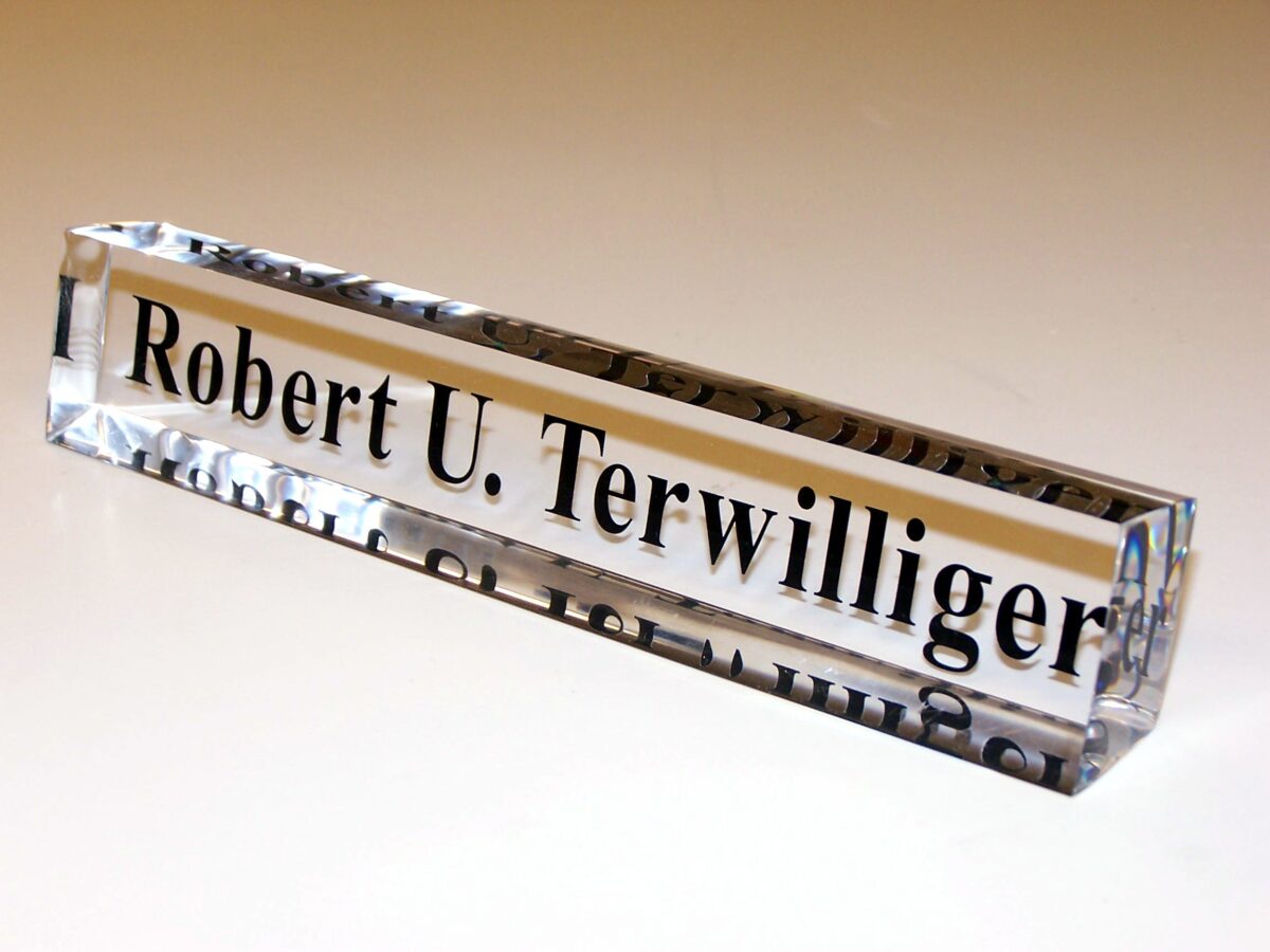 A rectangular glass nameplate with black text that reads "Robert U. Terwilliger" is displayed against a light background. The transparent glass reflects light, highlighting the crisp, clear text within.
