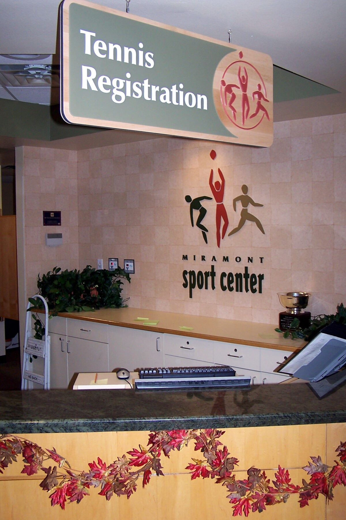 A tennis registration area at a sports center. The desk has a decorative fall-themed garland and a sign above reads "Tennis Registration." The wall behind the desk features the sports center's logo with vibrant figures and the name "Visionsport Sport Center.