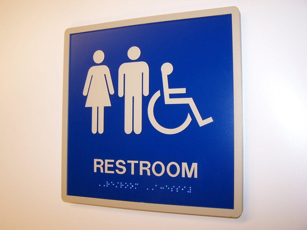 A blue restroom sign featuring white symbols for male, female, and wheelchair accessibility. The word "RESTROOM" is printed below the symbols, with additional text in Braille beneath it.