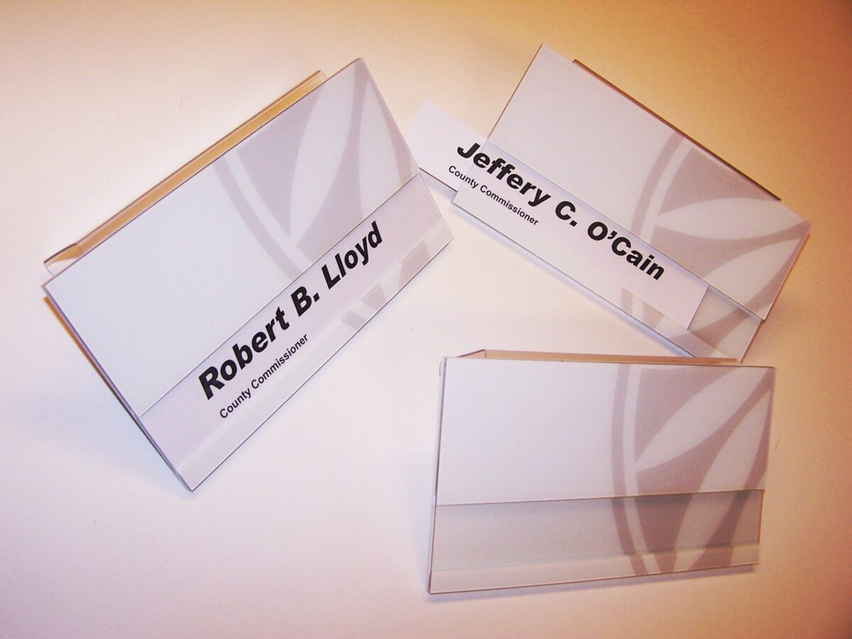Three nameplates are displayed against a white surface. Each nameplate is clear with black text showing names: "Robert B. Lloyd," "Jeffery C. O'Cain," and another with the name blank. The nameplates are positioned upright in holders.