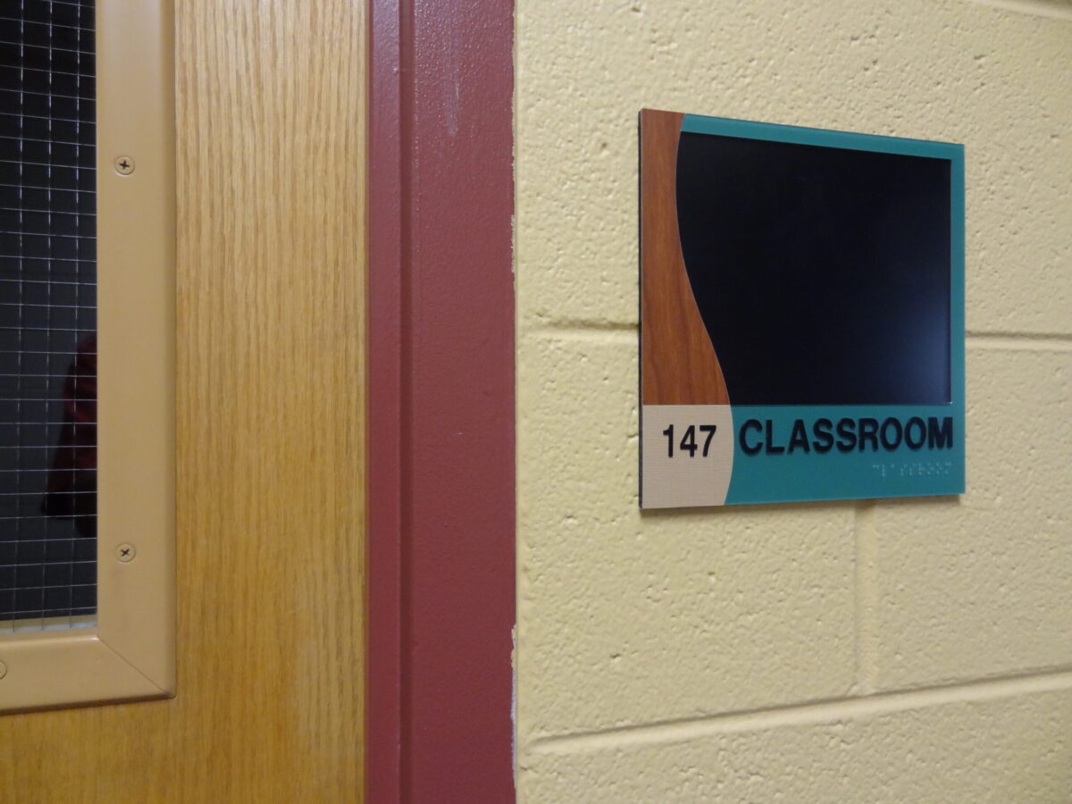 A door with a window panel on the left side and a classroom sign on the right. The sign is marked "147 CLASSROOM" with a decorative black and wood-colored design at the top.