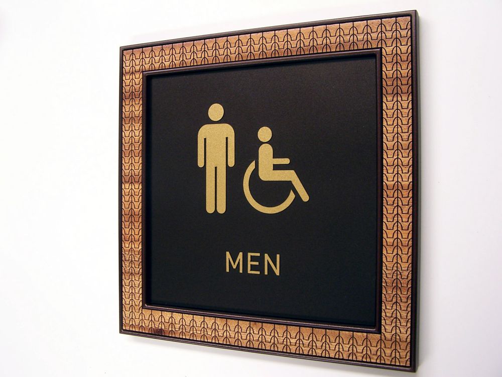 A bathroom sign indicating a men's restroom, featuring gold icons of a standing man and a person in a wheelchair on a black background. The sign is surrounded by a decorative, patterned frame.