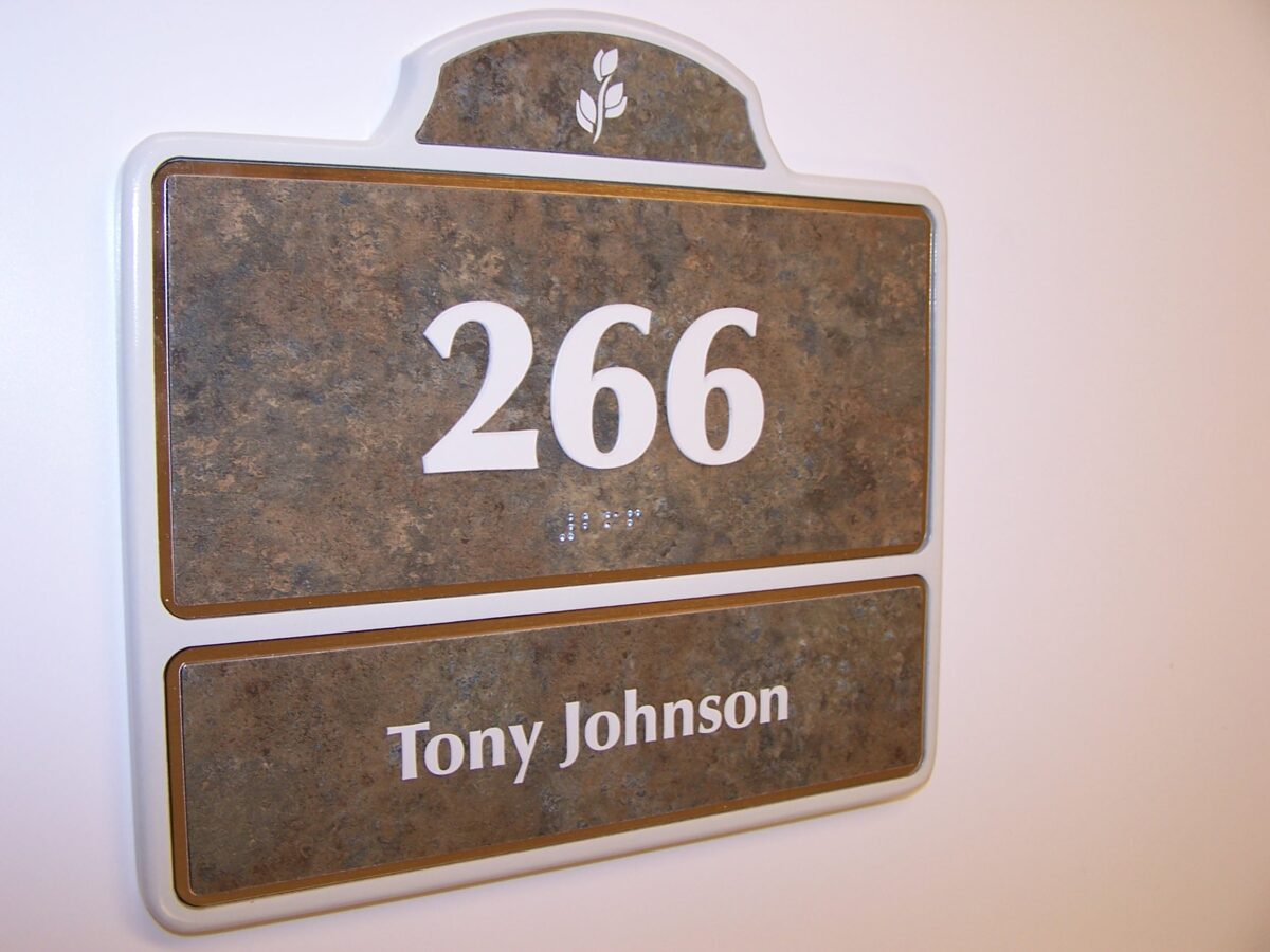 A room sign with a stone-like texture displays the number 266 in large white text. Below the number, in smaller white text, is the name Tony Johnson. The sign is mounted on a white wall.
