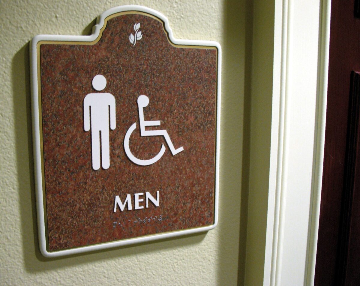 A restroom sign on a wall indicating a men's restroom. The sign features icons of a male figure and a person in a wheelchair, along with the word "MEN" beneath them. The sign has a brown background with white text and border.