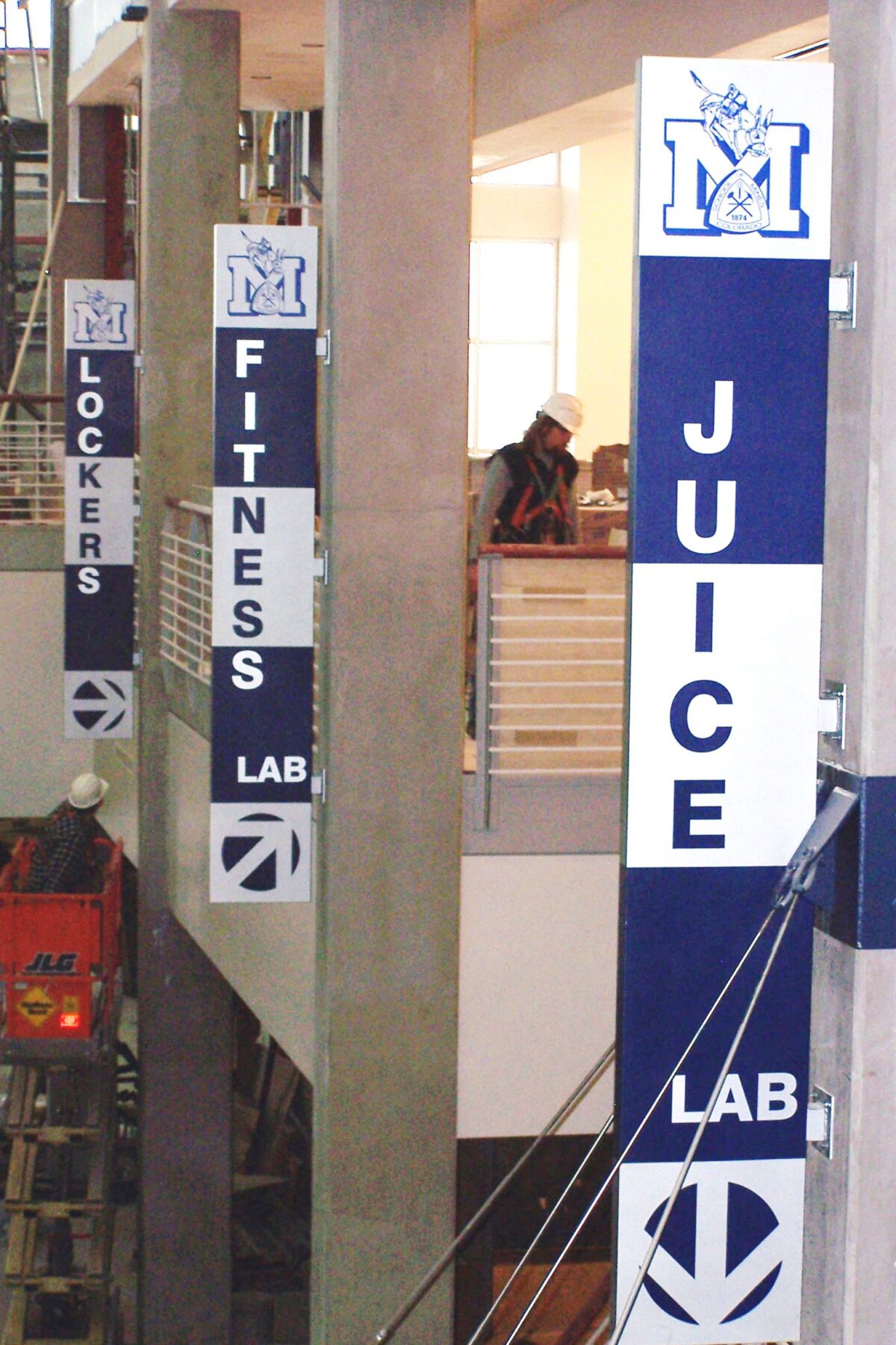 Banners hang vertically from columns inside a multi-level building, labeled "LOCKERS," "FITNESS LAB," and "JUICE LAB," respectively. Each banner features the same blue and white color scheme, with the letter "M" and a stylized knight's helmet logo at the top.