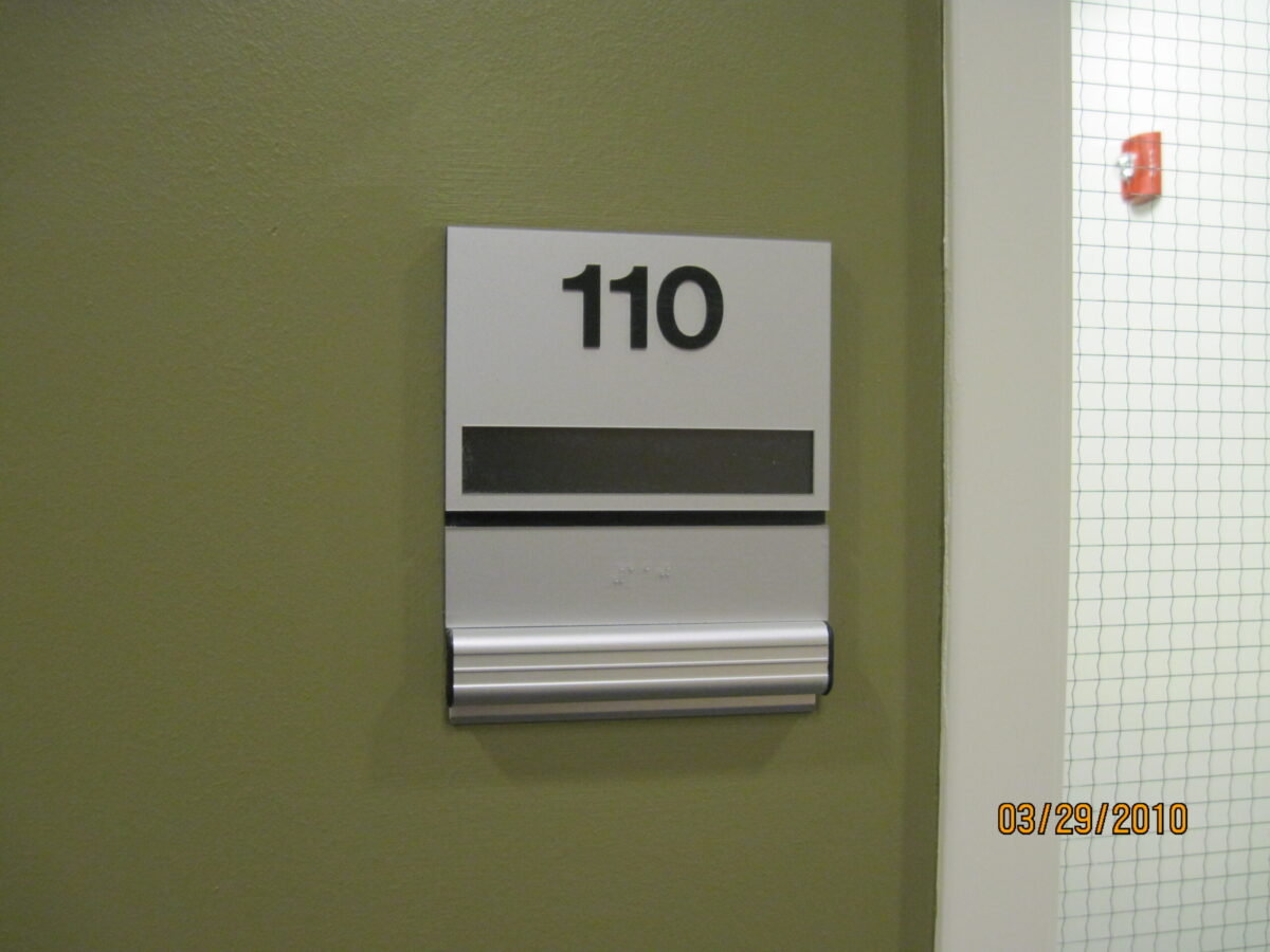 A sign on a wall displays the number "110" in bold black font. Below the number, there is a blank black stripe and a horizontal strip with braille. A partial white door frame with a mesh window is visible on the right.