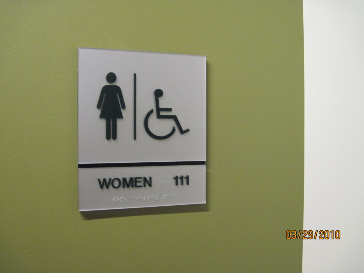 A sign on a green wall indicating a women's restroom with accessibility features. The sign includes icons of a woman and a wheelchair, the word "WOMEN," and the number "111" as well as braille text underneath.