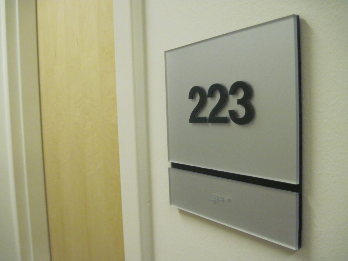 Sign displaying room number 223 mounted on a wall near a light wooden door in a building hallway. The sign has a metallic appearance with bold black numbers and a narrow horizontal strip underneath.