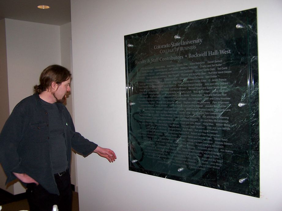 A person with long hair wearing a dark jacket and shirt points to a polished stone plaque mounted on a white wall, which appears to contain inscribed names or text.