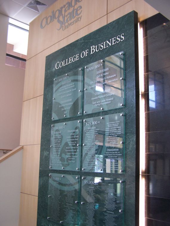 An interior display at a university's College of Business features a tall, green marble-like plaque with glass panels listing names. The backdrop includes wooden paneling and part of a stairwell. Natural light illuminates the area.