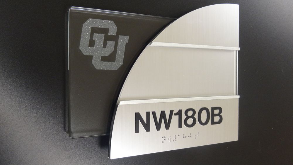 A wall-mounted nameplate with a sleek design features the text "NW180B." The background includes a dark gray panel with an emblem consisting of the letters "CU" intertwined. The nameplate itself is silver with a partial circular cutout on the left side.