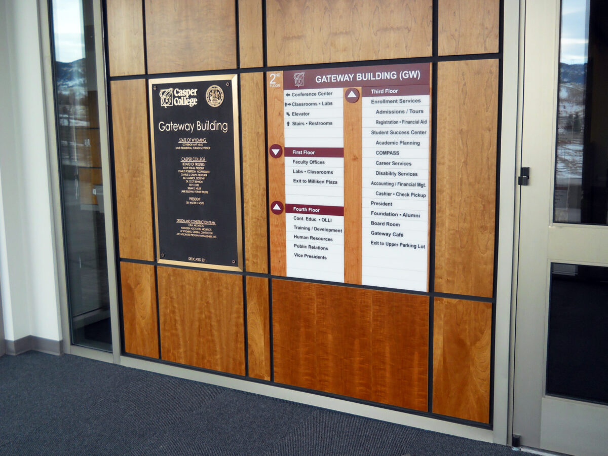 Wooden wall with two signage panels. The left panel lists various organizations and institutions; the right panel is a directory with floors and names of services and departments. A partially visible door is to the right. Tan and brown color scheme.