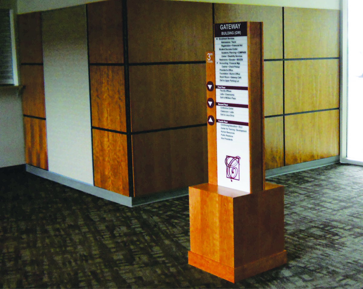 A wooden directory sign stands in the corner of a carpeted hallway with wood-paneled walls. The sign displays information and directional arrows. The area appears well-lit with natural and artificial light.