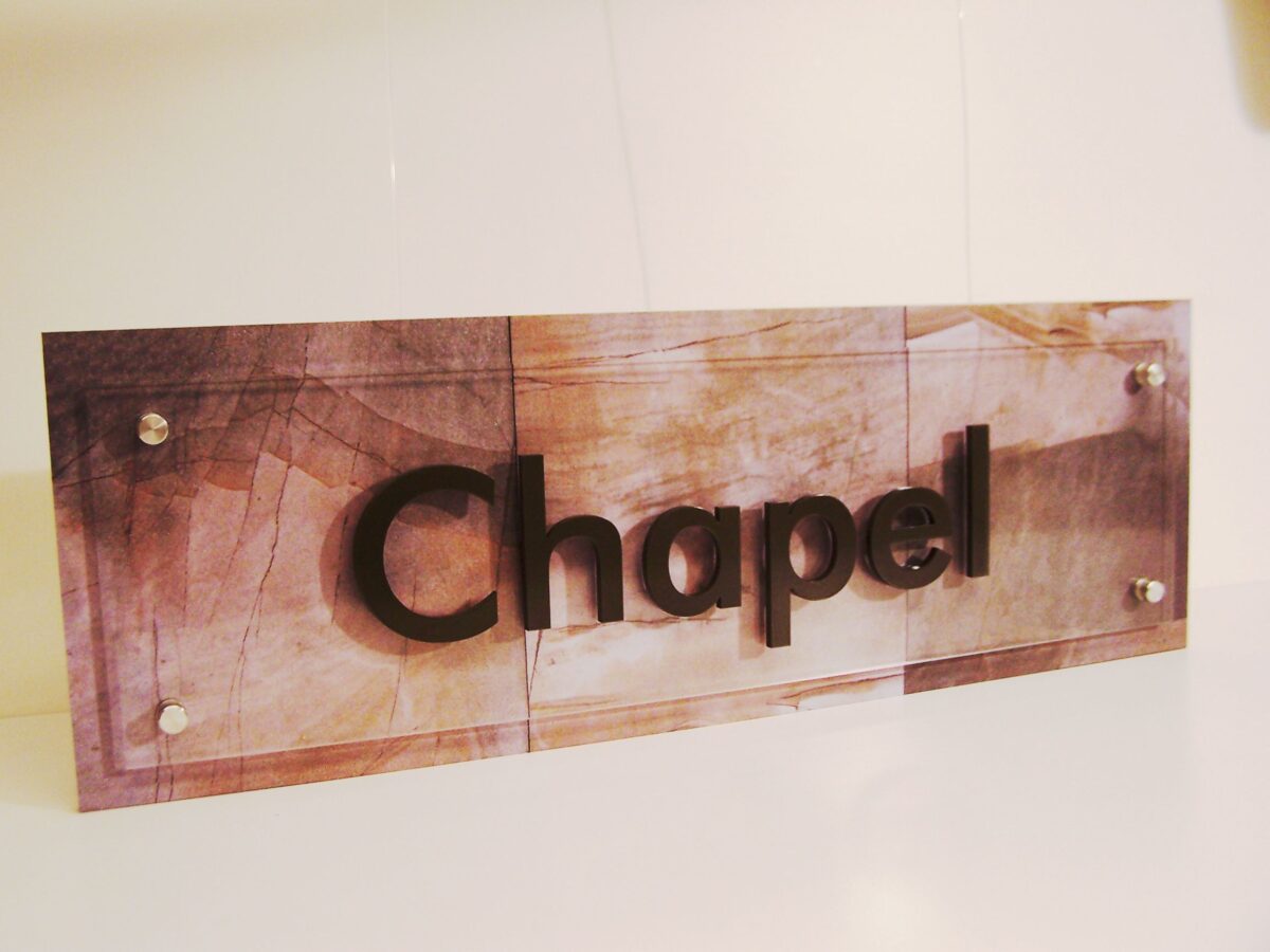 A sign mounted on a wall reads "Chapel" in bold, raised lettering. The background of the sign has a subtle, earthy-toned pattern with a rectangular shape and secured at the corners with visible screws. The overall look is modern and clean.
