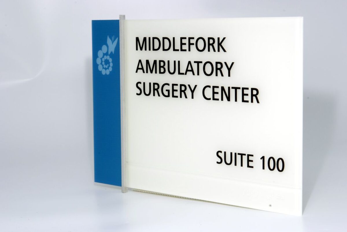 A sign for "Middlefork Ambulatory Surgery Center, Suite 100" against a white background. The sign is mounted on a base with a blue vertical stripe on the left side, featuring a small white emblem.