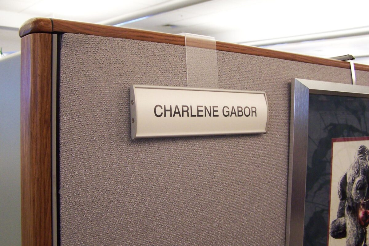 A nameplate reading "CHARLENE GABOR" is affixed to a cubicle wall. The surroundings include a grey fabric partition and part of a picture frame on the right side. The nameplate is held up by a clear hanger over the edge of the cubicle wall.