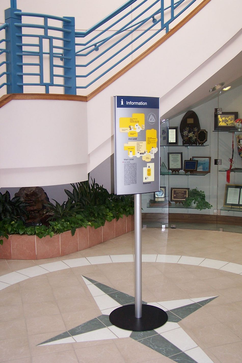 A stand-alone information board with yellow and white details is positioned in the center of an indoor area with tiled flooring and a compass design.