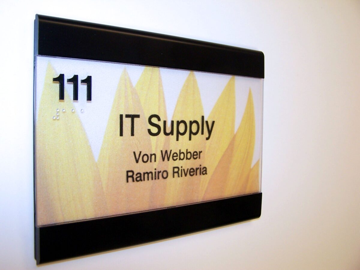 A door sign with a sunflower design shows the text "111 IT Supply" and lists two names: "Von Webber" and "Ramiro Riveria." The sign is mounted on a white wall.
