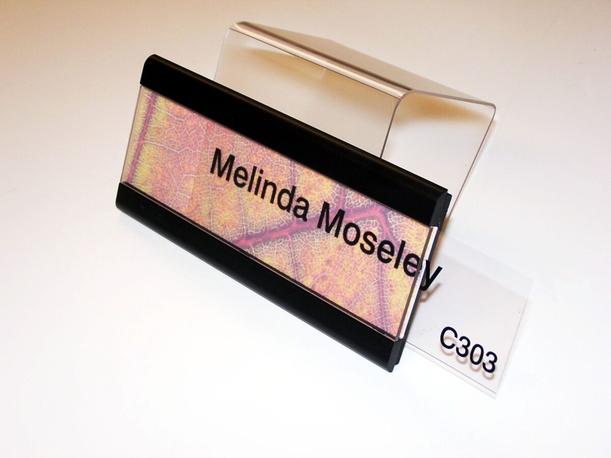 A nameplate with the text "Melinda Moseley" is displayed on a clear stand. The nameplate has a multicolored background with a mix of pink, yellow, and purple hues, and is bordered by black strips at the top and bottom. The label "C303" is visible beneath the stand.