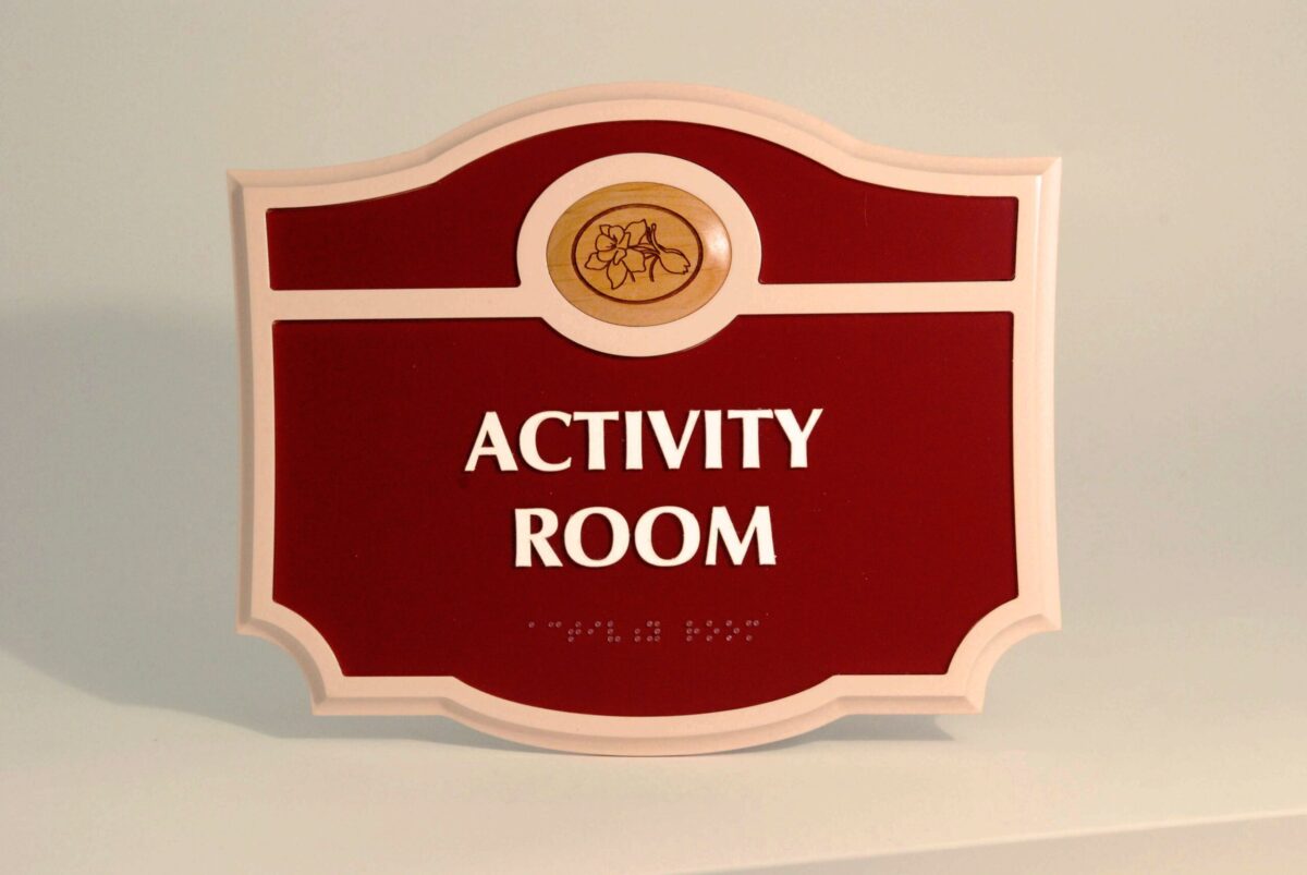 A red and white sign with an ornate shape, displaying the words "ACTIVITY ROOM" in bold white letters. The top part of the sign features a small circular emblem with an illustration of a flower inside it.