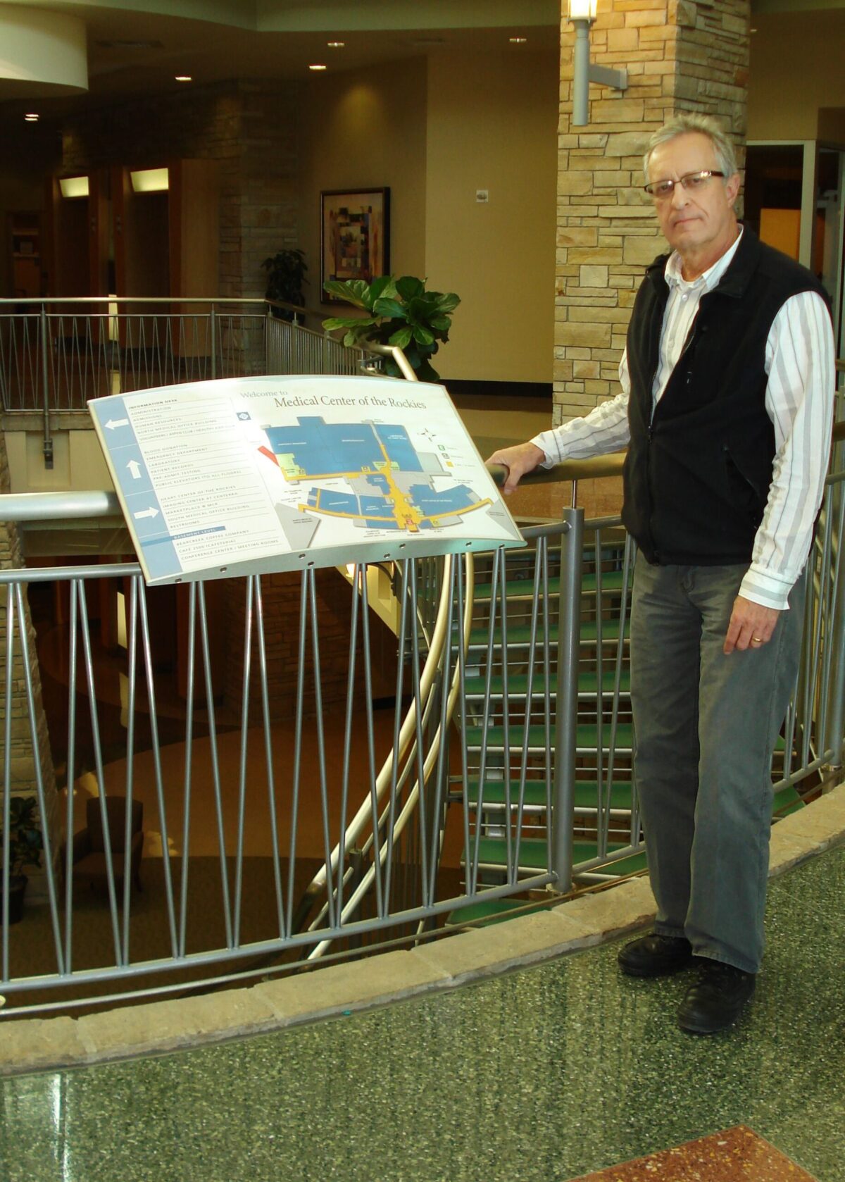 A person standing indoors next to a large directory sign mounted on a railing. The directory displays a map and text. The person is dressed in a striped shirt, dark vest, gray pants, and black shoes, and is looking towards the camera. The background includes a staircase.