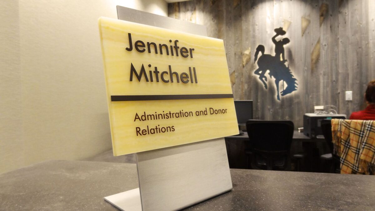 A desktop nameplate reads "Jennifer Mitchell, Administration and Donor Relations." The background shows a blurred office space with a person sitting at a desk and a wall decoration featuring a silhouette of a cowboy on a bucking horse.