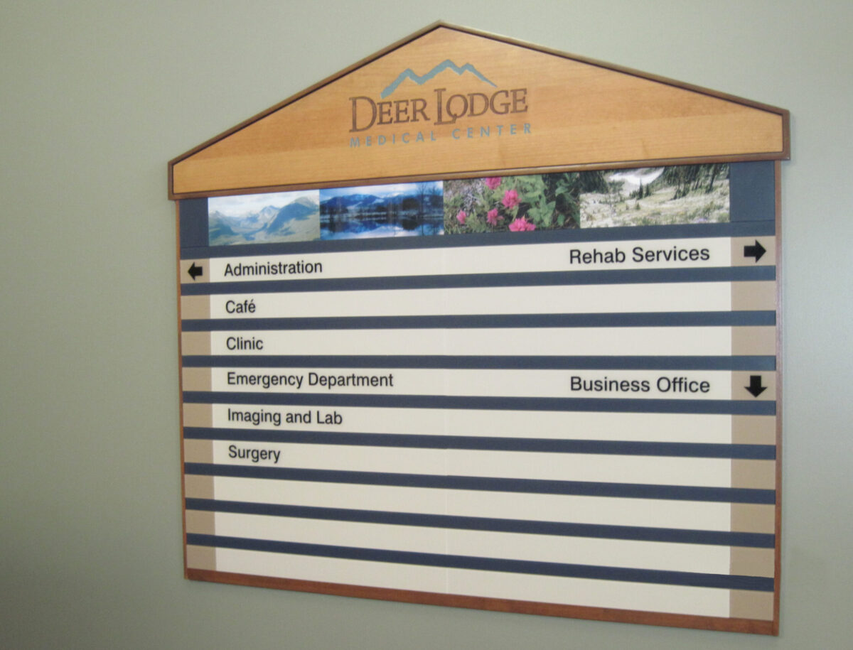 A wooden directory sign mounted on a wall, displaying various departments at Deer Lodge. Categories listed include Administration, Calls, Clinic, Emergency Department, Imaging and Lab, Surgery, Rehab Services, and Business Office, with arrows indicating directions.