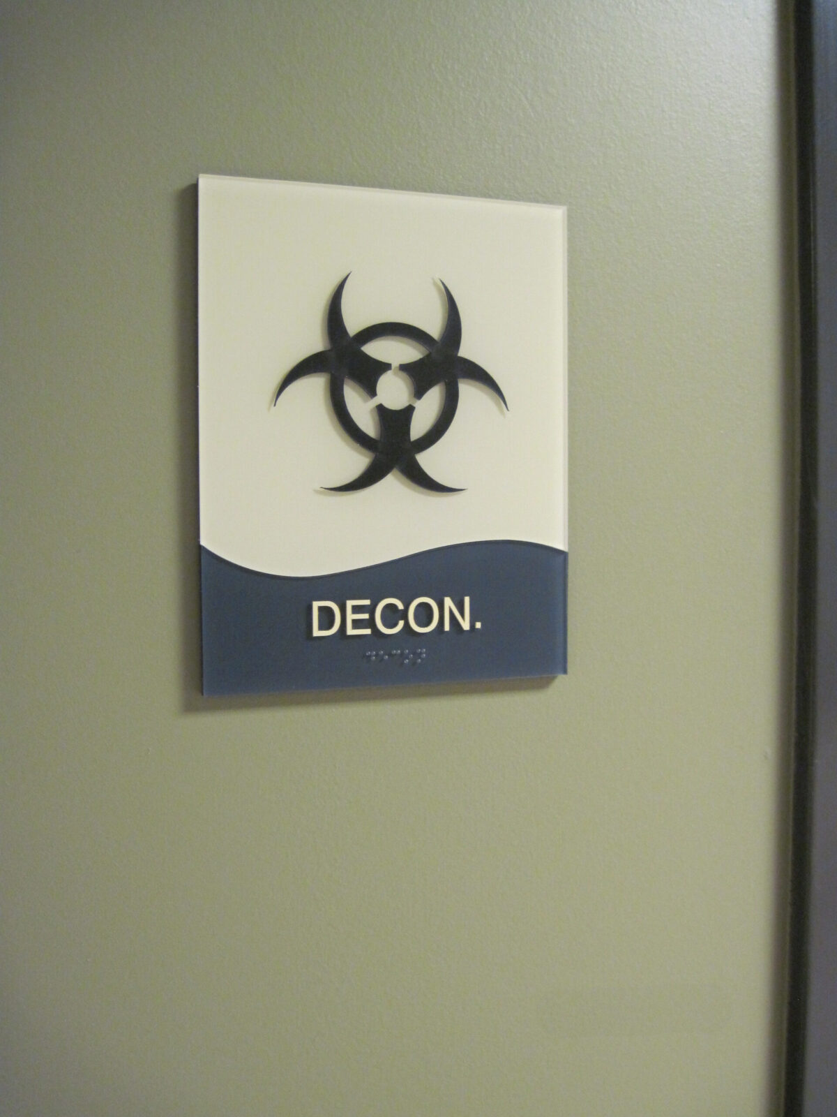 A sign on a wall has a black biohazard symbol above the word "DECON." The background is divided into an upper white section and a lower gray section.