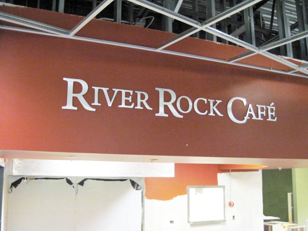 A sign for "River Rock Café" is displayed on a red-brown wall with white letters. The surrounding area appears to be under construction, with exposed ceiling beams and an unfinished section below the sign.