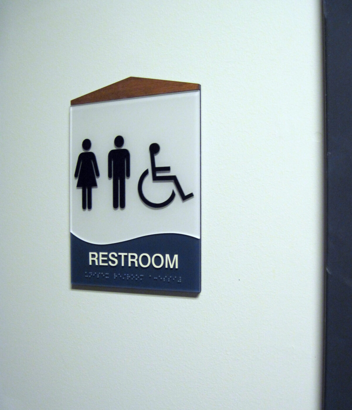 A restroom sign on a wall features symbols for women, men, and wheelchair accessibility above the word "Restroom." The top of the sign has a brown triangular accent.
