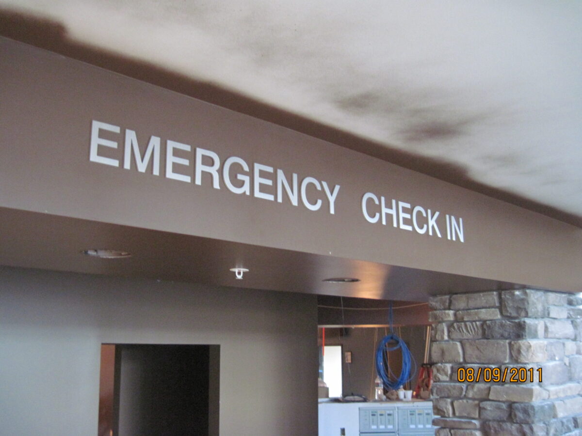 Signage inside a building reads "EMERGENCY CHECK IN" in bold white letters on a dark brown beam. The date on the bottom right corner shows 08/19/2011. The area underneath appears to be part of a reception or waiting area, with a stone wall visible in the background.