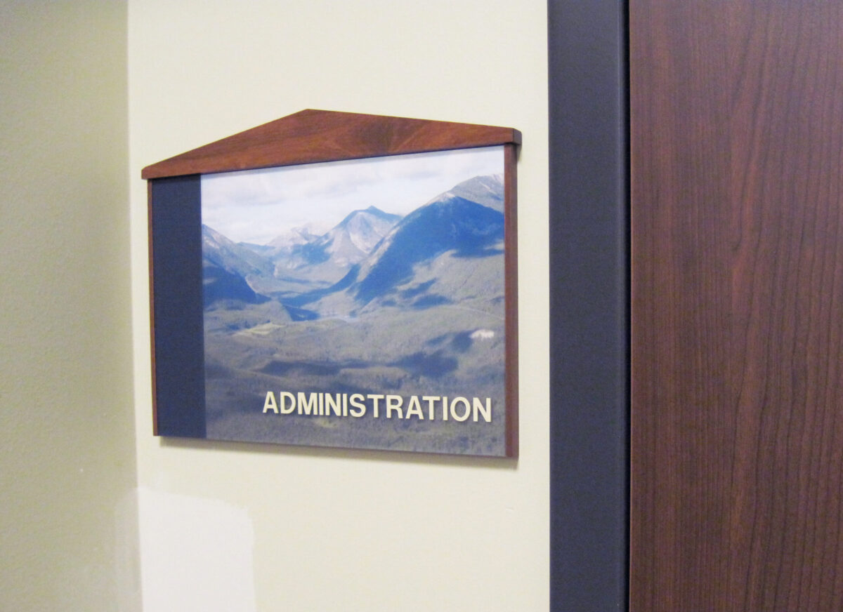A wall sign for an administration office features a photograph of mountainous scenery beneath a wooden frame with a peak at the top. The word "ADMINISTRATION" is written in capital letters at the bottom of the image. A wooden door is partially visible beside the sign.