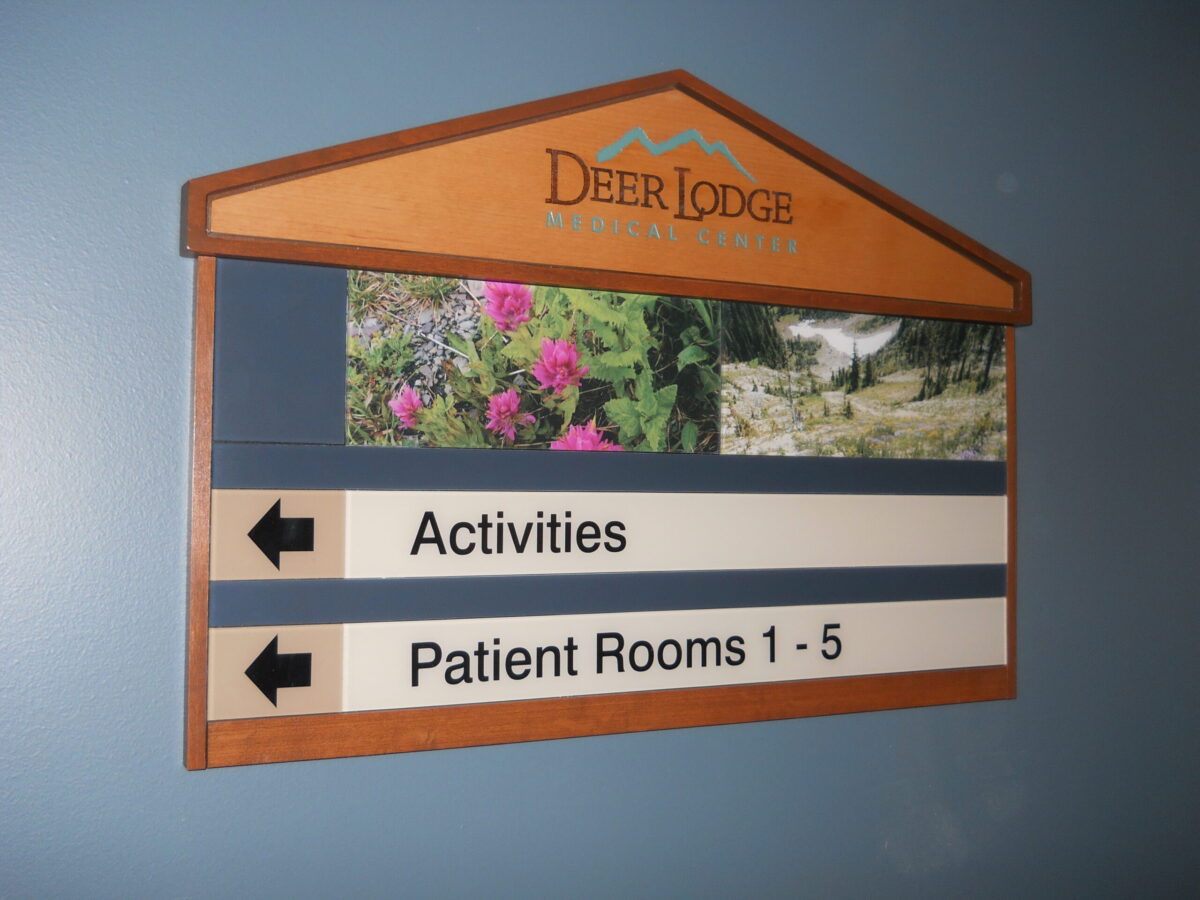 A wall-mounted directional sign with a wooden frame, labeled "Deer Lodge" at the top. Below are arrows pointing left for "Activities" and right for "Patient Rooms 1-5." The sign features small images of flowers and a nature scene.