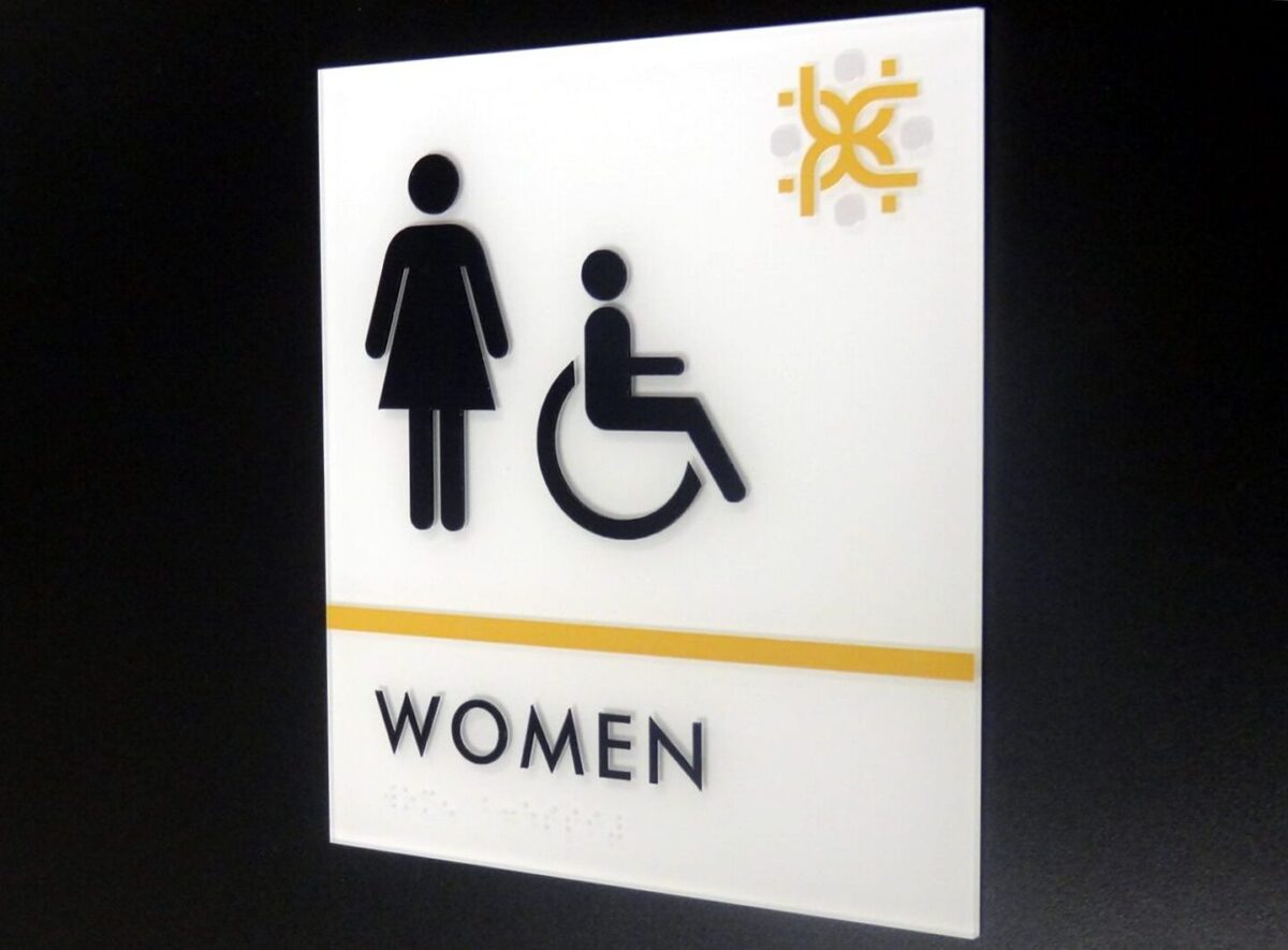 A sign with symbols indicating a women's restroom. It includes a pictogram of a woman and a wheelchair user. The word "WOMEN" is written in black text below the symbols, with a yellow line above the text. The upper right corner features a decorative logo.