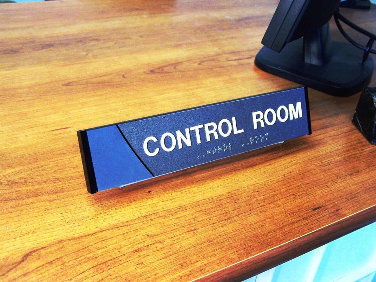 A small rectangular sign on a wooden surface reads "CONTROL ROOM" in bold white letters with Braille text below. The sign has a sleek black and blue color scheme, and a computer monitor is visible in the background.