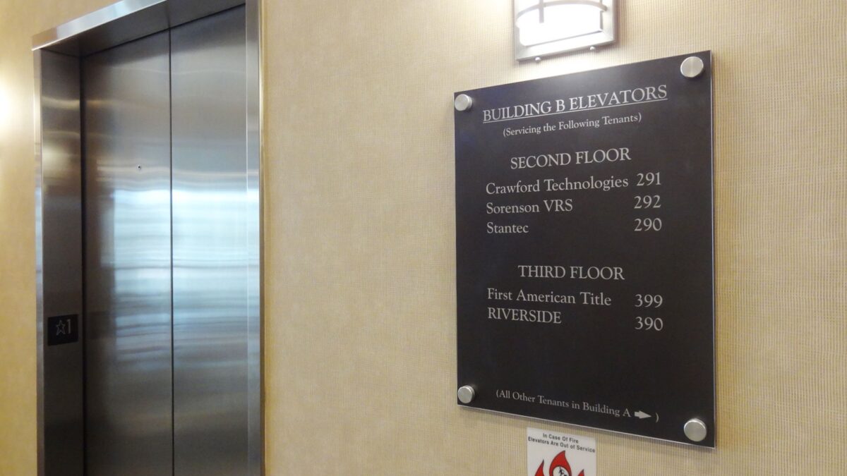 A directory board is mounted on a beige wall next to an elevator. It lists the names and room numbers of businesses on the second and third floors, such as Certified Technologies in room 201 and First American Title in room 300.