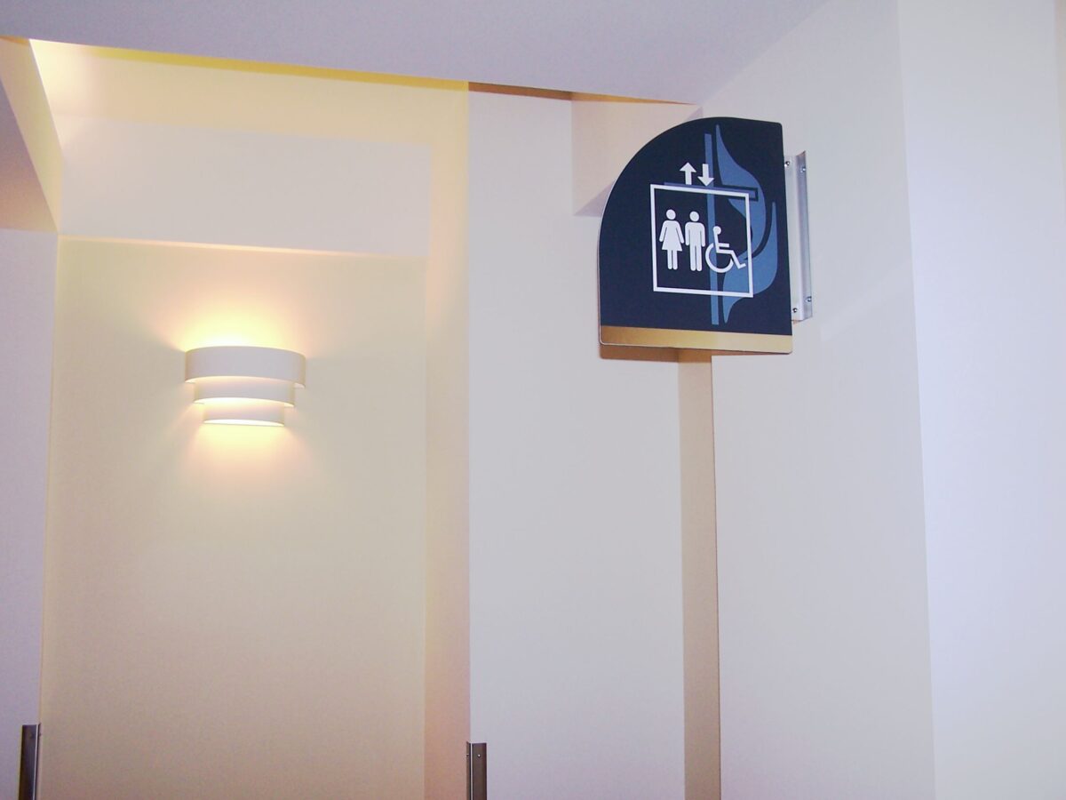 A restroom sign mounted on a wall, indicating male, female, and wheelchair accessible facilities. The background is a softly lit, light-colored wall with a modern sconce light fixture.