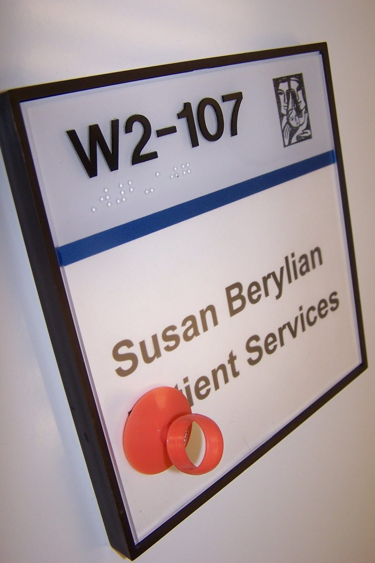 A room sign with the text "W2-107" and "Susan Berylian Client Services" is mounted on a wall. The sign has a blue stripe and a small icon in the top right corner. Braille text is above the blue stripe.