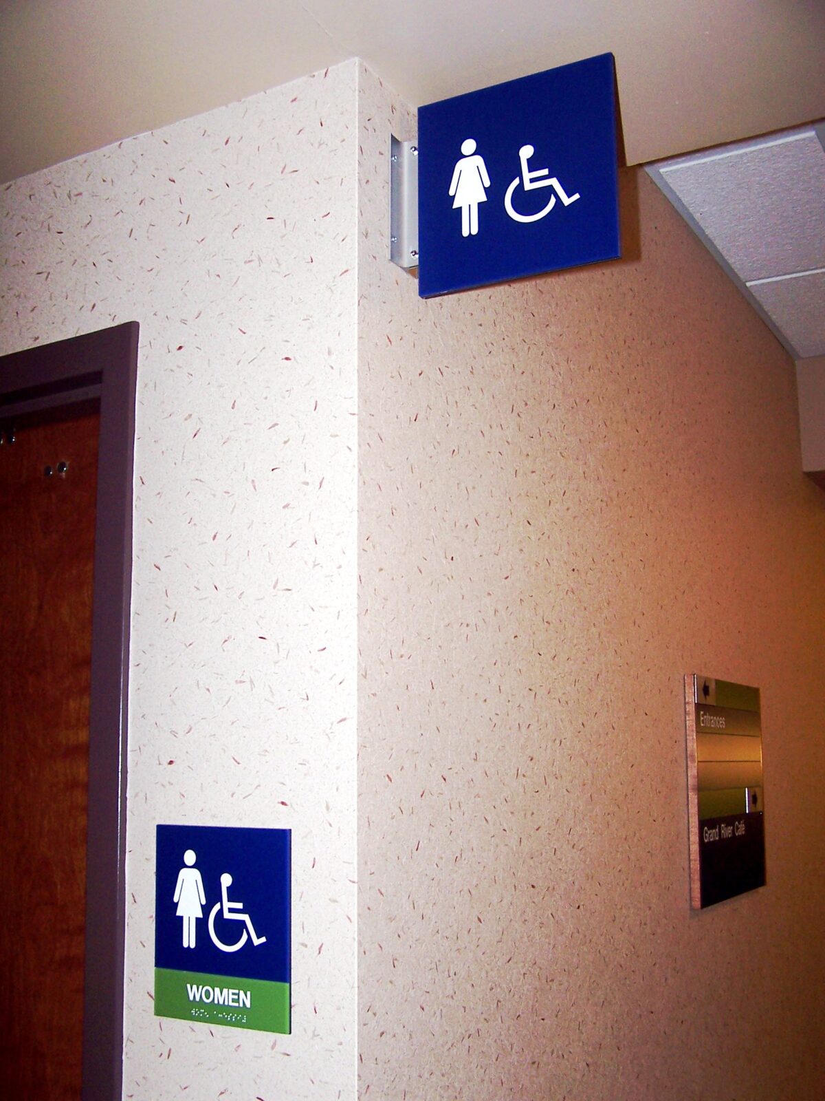 A corner in a building hallway shows a wall-mounted sign indicating restrooms for women and those with disabilities. Below, another similar sign is positioned on the wall next to the restroom door. Both signs feature accessible icons.