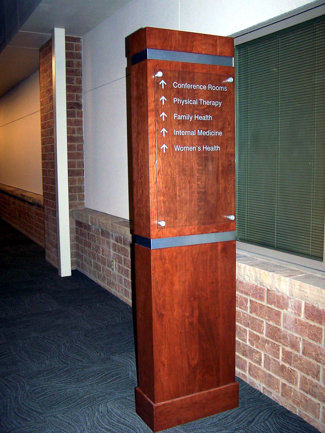 A wooden directional signpost in a hallway points to different locations including conference rooms, physical therapy, family health, internal medicine, and women's health. The sign features white text and arrows on a wood panel set against a brick wall and carpeted floor.
