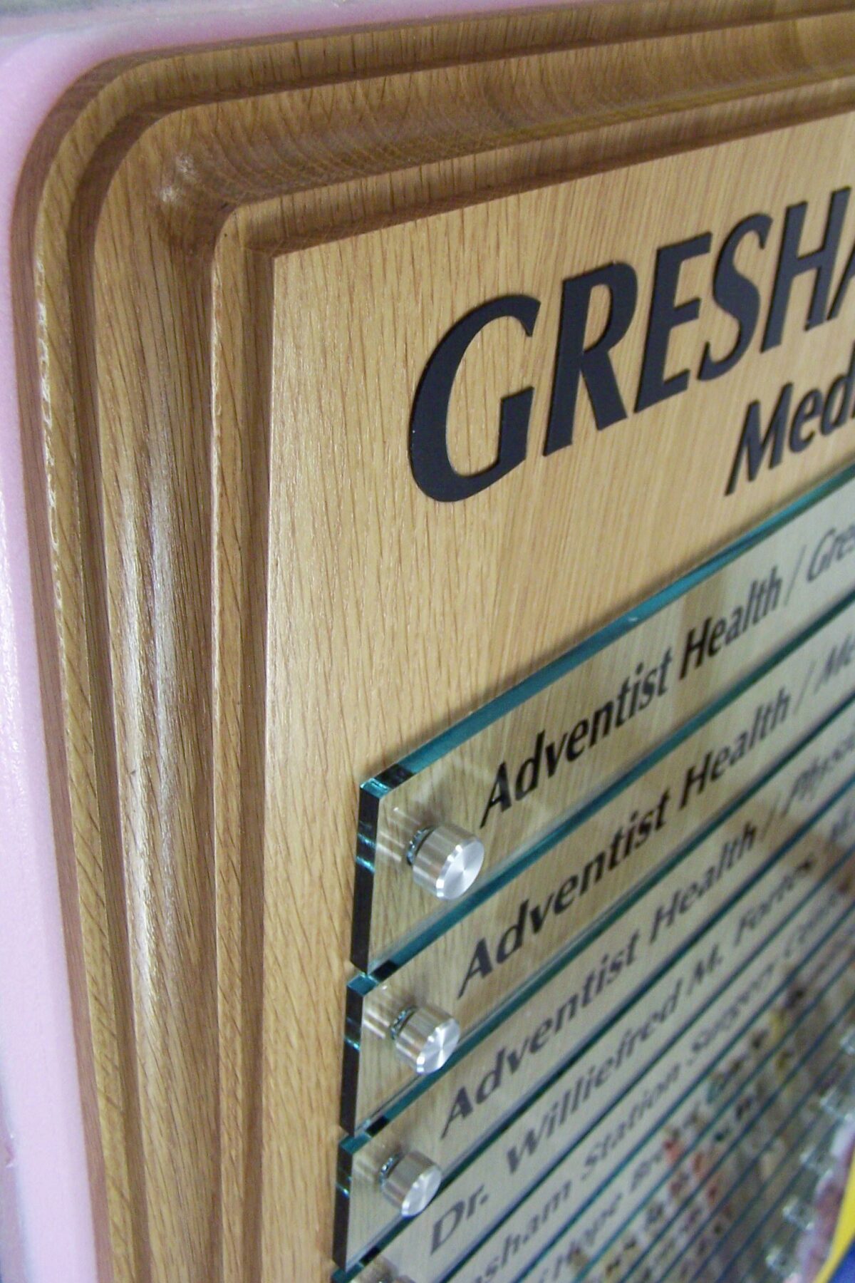 A close-up shot of a wooden directory sign with glass panels on a wall. It has several sections listing different names and titles. The partial text "GRESHAM" is visible at the top. The layers of the sign create a three-dimensional effect.