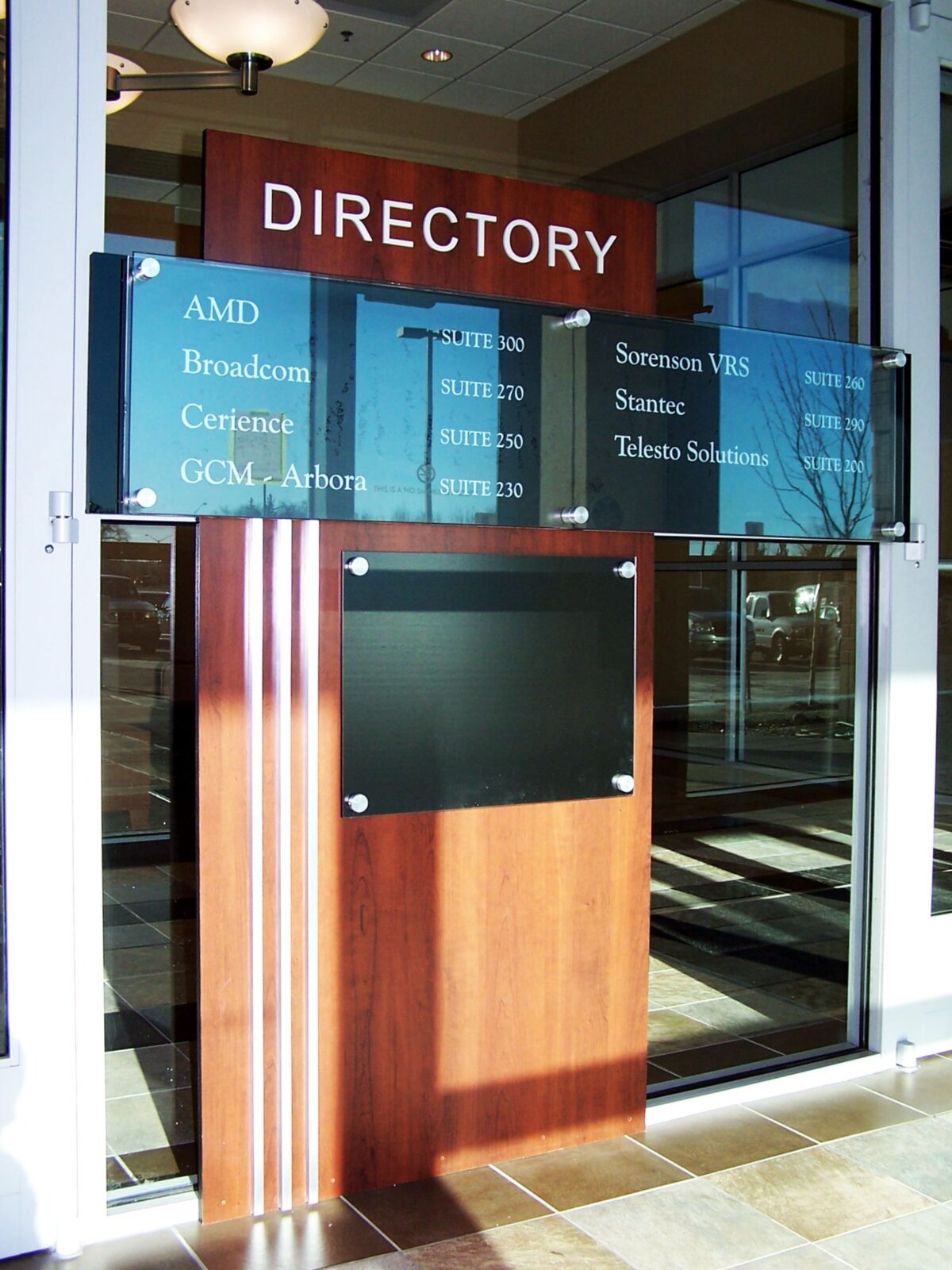 A directory sign mounted on a wooden board in a lobby area. The board lists various company names such as AMD, Broadcom, and others, with corresponding suite numbers. Below the listings is a blank black space for additional information. Glass doors are visible in the background.