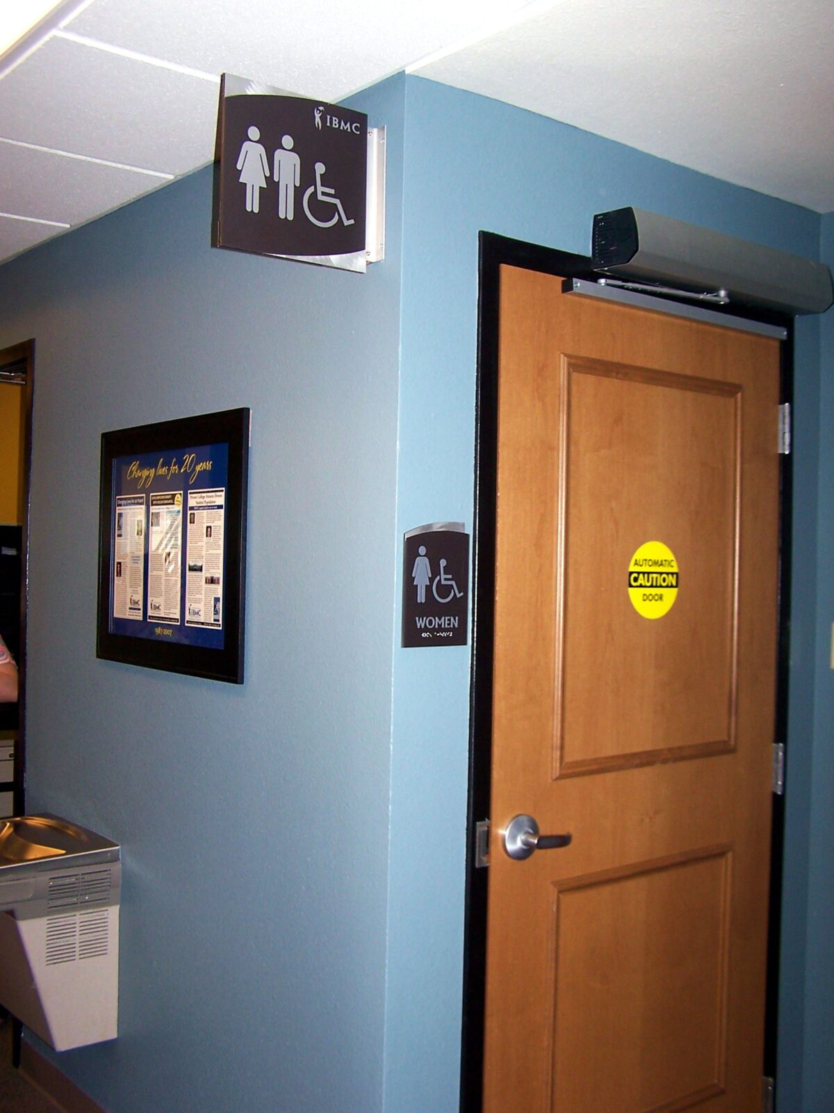 A restroom door with a wooden finish is shown. Above the door is a sign indicating it is a unisex and accessible restroom. A wall-mounted sign adjacent to the door repeats the same information. A yellow caution sticker is on the door, which is closed.